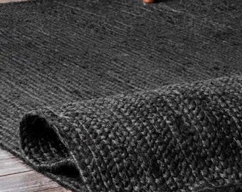 Könnte beinhalten: Ein aufgerollter, dunkelgrauer gewebter Teppich. Der Teppich besteht aus einem dicken, strukturierten Material und ist teilweise aufgerollt, wodurch die Unterseite sichtbar wird. Der Teppich liegt auf einem Holzboden, im Hintergrund sind ein Schrank und ein Stuhl zu sehen.
