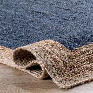 Op de afbeelding: Close-up van een opgerolde blauwe en beige jute vloerkleed. Het vloerkleed heeft een geweven textuur met een verloop van blauwe tinten, overgaand in een natuurlijke beige rand. Het vloerkleed is gemaakt van natuurlijke vezels en is geschikt voor binnengebruik.