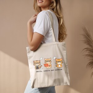 May include: Beige tote bag featuring three cartoon cats wearing glasses, each with a different accounting tool: calculator, laptop, and coffee cup. The text "debits, credits and caffeine" is printed below the cats. The bag has cream-colored handles.