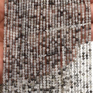 以下が含まれることがあります： グレーとクリアカラーの小さな丸いビーズの複数のストランドのクローズアップ。ビーズは手で持たれており、繊細な質感が示されています。これらのビーズは、おそらくジュエリー作りに使用されます。