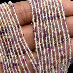 Op de afbeelding: Meerdere strengen kleine, ronde kralen in tinten paars, geel en helder. De kralen zijn aan elkaar geregen, waardoor een delicate en kleurrijke weergave ontstaat. De kralen worden in een hand gehouden tegen een zwarte achtergrond.