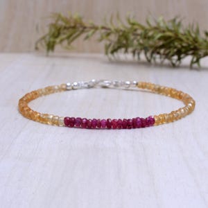 May include: A delicate bracelet featuring a combination of small, round gemstones. The bracelet has a gradient of colors, transitioning from a golden yellow to a vibrant red. The clasp is silver. The bracelet is displayed on a light-colored surface.