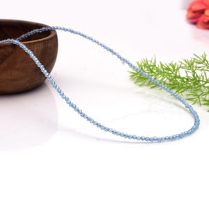 May include: A fine necklace composed of tiny, pale blue beads. The beads are strung closely together, creating a gentle shimmer. The necklace is shown on a white surface, with a wooden bowl and foliage in the background.