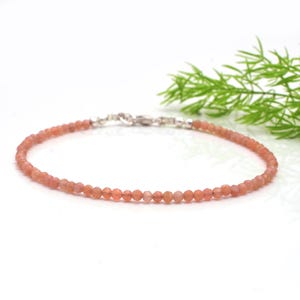 Peut inclure: Bracelet délicat composé de petites perles rondes de couleur pêche. Les perles sont enfilées et fixées par un fermoir argenté. Le bracelet est présenté sur fond blanc, avec un peu de feuillage vert.