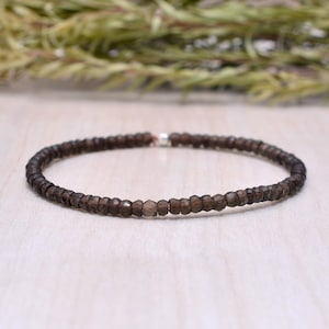 Puede incluir: Una pulsera de cuentas marrón oscuro. La pulsera está hecha de pequeñas cuentas facetadas ensartadas. Las cuentas son una mezcla de tonos marrones y grises. La pulsera se exhibe sobre una superficie de madera clara.