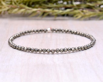 May include: A delicate bracelet composed of small, round, metallic beads. The beads have a faceted appearance, reflecting light. The bracelet is a neutral silver color and appears to be strung on a thin, clear elastic cord. The background is a light wood surface.