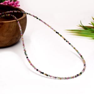 Può includere: Una delicata collana composta da piccole perline di pietre preziose multicolori. Le perline mostrano una gamma di colori, tra cui rosa, verde, giallo e nero. La collana è esposta su una superficie bianca, con una ciotola di legno e verde sullo sfondo.