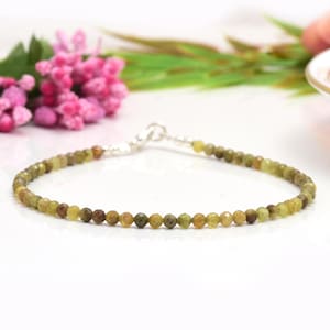 Pode incluir: Uma pulseira delicada com pequenas contas facetadas em tons de verde oliva e castanho. As contas são enfiadas com um fecho prateado. A pulseira é exibida em uma superfície branca, com flores cor-de-rosa e folhas verdes ao fundo.