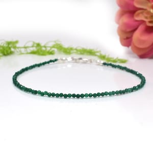 Könnte beinhalten: Ein zartes, dunkelgrünes Armband oder Fußkettchen mit Perlen. Die kleinen, runden Perlen sind mit einem silbernen Verschluss aufgefädelt. Der Schmuck wird auf einer weißen Oberfläche mit einer rosa Blume und grünem Laub im Hintergrund präsentiert.