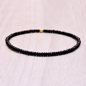 Puede incluir: Una delicada pulsera de cuentas negras con una cuenta dorada como acento. La pulsera está hecha de pequeñas cuentas negras facetadas ensartadas. La cuenta dorada está centrada en la pulsera. Se muestra sobre una superficie de madera clara.