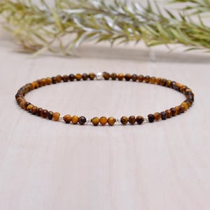 May include: A beaded bracelet or anklet featuring small, faceted tiger eye stones in shades of brown and gold. The bracelet is strung on a flexible cord and secured with a silver clasp. The jewelry is displayed on a light-colored surface.
