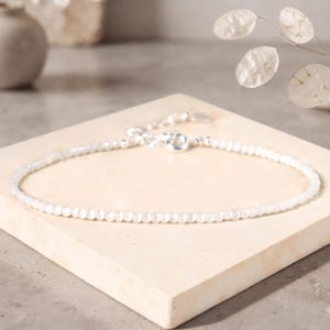 Può includere: Una delicata cavigliera composta da un singolo filo di piccole perline bianche iridescenti. Il gioiello presenta una chiusura in metallo color argento e una catenella di estensione regolabile, offrendo una lucentezza sottile e raffinata.