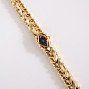 May include: A gold-tone watch featuring a textured, chevron-patterned link bracelet and a marquise-shaped case. The watch face is black with a single small stone at the twelve o'clock position and gold-coloured hands for hour and minute tracking.