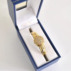 May include: A gold-toned hinged bangle bracelet featuring a circular centre piece encrusted with numerous small clear stones, set within a deep blue rectangular presentation box with a white interior lining and a hinged lid.