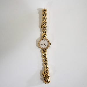 May include: A gold-tone wristwatch featuring a round white dial with Roman numeral markers and small clear stones at the cardinal points. The watch band is composed of interconnected leaf-shaped gold-tone metal links with a secure clasp.