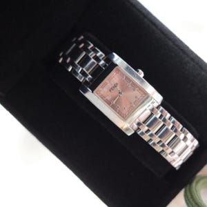 Può includere: Un orologio Fendi con quadrante rosa rettangolare e cinturino argentato. L'orologio è presentato su una superficie nera. Il quadrante dell'orologio ha il logo Fendi.