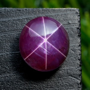Puede incluir: Una gema redonda y pulida, de color púrpura intenso, con un patrón de estrella de seis rayos. El efecto de estrella se crea por la luz que se refleja en las inclusiones dentro de la piedra. La gema está sobre una superficie oscura y texturizada.