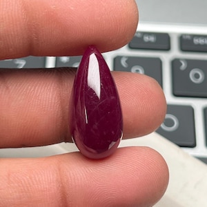 Puede incluir: Una gema de rubí pulida en forma de lágrima. La gema es de un color rojo intenso y rico con una superficie brillante. El rubí se sostiene entre dos dedos, con un teclado de ordenador en el fondo.