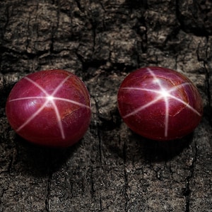 Pode incluir: Duas pedras preciosas de rubi estrela ovais, com um tom vermelho profundo e um padrão de estrela de seis raios. As gemas estão sobre um fundo escuro e texturizado. Ideal para a criação de joias.