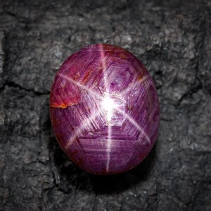 Puede incluir: Una gema de rubí estrella pulida, de forma ovalada, con un efecto de estrella de seis puntas. La gema es de color rojo púrpura intenso, con un patrón de estrella que irradia desde un punto central. Fondo oscuro y texturizado.