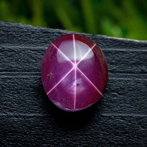 Puede incluir: Una gema pulida, de forma ovalada, de color rojo púrpura, con un patrón de estrella de seis rayos. La gema está colocada sobre una superficie oscura y texturizada, con un fondo verde borroso.