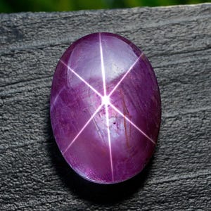 Puede incluir: Gema de rubí estrella ovalada de color púrpura intenso. La piedra pulida muestra un efecto de estrella de seis rayos, con líneas blancas brillantes que irradian desde un punto central. La gema está sobre una superficie oscura y texturizada.