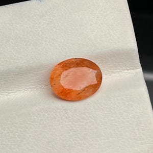 Puede incluir: Una gema ovalada de color naranja con un centro más claro, color melocotón. La gema está colocada sobre una superficie blanca texturizada, con un fondo oscuro visible en la esquina.