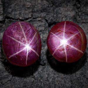 Op de afbeelding: Twee ovale sterrobijn edelstenen. De edelstenen hebben een diepe paarsrode kleur, met een zesstralig sterreneffect zichtbaar op hun gepolijste oppervlakken. De achtergrond is een donker, getextureerd oppervlak.