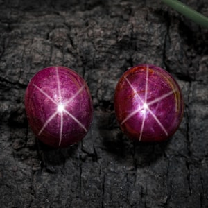 Puede incluir: Dos rubíes estrella ovalados de color rojo intenso, con un efecto de estrella de seis rayos. Las piedras preciosas se colocan sobre un fondo oscuro y texturizado, resaltando su color vibrante y su fenómeno óptico único. Joyería de piedras preciosas.