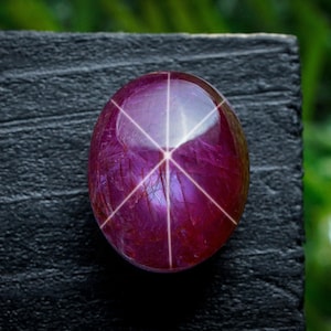 May include: An oval-shaped ruby gemstone, exhibiting a six-rayed star effect. The gem is a deep purplish-red hue, with white lines emanating from the centre. It is displayed on a dark, textured surface, with a blurred green backdrop.