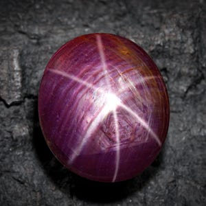 Puede incluir: Una gema de rubí estrella tallada en cabujón. La gema es de color rojo púrpura intenso con un efecto de estrella de seis rayos. La estrella es blanca y prominente sobre la superficie del rubí. La gema está sobre un fondo oscuro y texturizado.