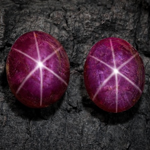 Könnte beinhalten: Zwei ovale, tiefrote Edelsteine mit einem sechsstrahligen Sternmuster. Die Edelsteine sind vor einem dunklen, strukturierten Hintergrund platziert, der ihre lebendige Farbe und den einzigartigen optischen Effekt hervorhebt. Wahrscheinlich handelt es sich um Sternrubine.