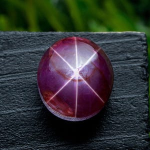 Op de afbeelding: Een gepolijste, cabochon-geslepen edelsteen met een stereffect. De edelsteen heeft een diepe paarsrode kleur, met een zesstralig sterpatroon dat vanuit het midden uitstraalt. De steen ligt op een donkere, getextureerde ondergrond.