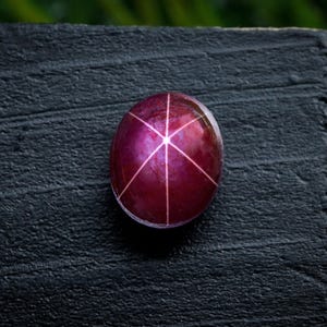 Puede incluir: Una gema pulida, de forma ovalada, de color rojo intenso, con un patrón de estrella de seis rayos. El efecto de estrella se crea por la luz que se refleja en las inclusiones dentro de la piedra. La gema está sobre una superficie oscura y texturizada.