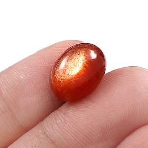 May include: An oval-shaped sunstone gemstone with a smooth, polished cabochon surface. The stone displays a deep reddish-orange base colour with a distinct, shimmering metallic golden aventurescence effect concentrated across the upper portion.