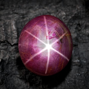 Może przedstawiać: Okrągły, wypolerowany kamień szlachetny z rubinu gwiazdowego. Klejnot ma głęboki, purpurowy kolor z sześcioramiennym wzorem gwiazdy wychodzącym z centralnego punktu. Kamień jest osadzony na ciemnym, teksturowanym tle.