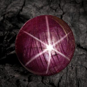 Puede incluir: Una gema redonda y pulida, de color rojo intenso, que muestra un efecto de estrella de seis rayos. El patrón de estrella emana de un punto central, creando un contraste visual llamativo contra la superficie de la piedra. La gema está sobre un fondo oscuro y texturizado.