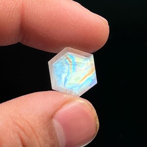 May include: A hexagonal gemstone with multiple facets, held between fingers. The gem exhibits a play of colours, including blues, yellows, and oranges, reflecting light. The stone has a polished, translucent appearance, with sharp edges. The background is black.