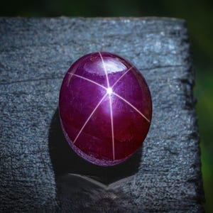 Puede incluir: Una gema pulida, de forma ovalada, de color púrpura oscuro, con un efecto de estrella de seis rayos. El patrón de estrella se crea mediante líneas blancas que se cruzan en la superficie de la gema. La gema está sobre un fondo oscuro y texturizado.