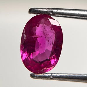 Może przedstawiać: Owalny, szlifowany kamień szlachetny w kolorze żywego różu magenta, trzymany przez srebrne metalowe pęsety. Fasetowany kamień wykazuje wewnętrzne cechy przejrzystości oraz odbicia światła na polerowanej powierzchni, prezentując głęboki, nasycony kolor.