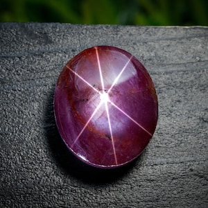 May include: A polished, oval gemstone in a deep burgundy colour. It displays a six-rayed star effect, with bright white lines emanating from the centre. The stone is set against a dark, textured surface, providing a strong contrast.