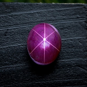 Puede incluir: Una gema pulida, de forma ovalada, de color magenta intenso, con un patrón de estrella de seis rayos. La gema está colocada sobre una superficie oscura y texturizada, con un fondo verde borroso.