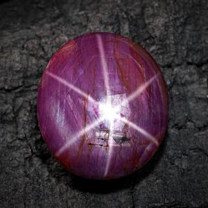 Puede incluir: Una gema de rubí estrella pulida, de forma redonda. La gema es de un color rojo púrpura intenso, que muestra un efecto de estrella de seis rayos que emana del centro. La piedra está sobre un fondo oscuro y texturizado.