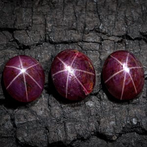 May include: Three oval-shaped, star rubies with a deep red-purple hue are displayed against a dark, textured background. Each gem features a six-rayed star pattern, with white lines radiating from a central point.