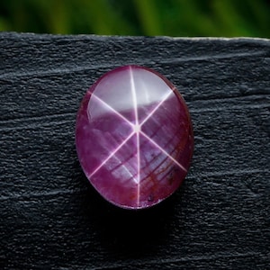 Peut inclure: Pierre précieuse rubis ovale polie, avec un effet d'étoile à six rayons. La gemme est d'un rouge-violet profond, avec un motif d'étoile blanc brillant rayonnant du centre. La pierre est posée sur une surface sombre et texturée.