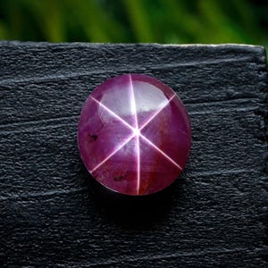 Puede incluir: Una gema de rubí estrella pulida y redonda, con un patrón de estrella de seis rayos. La gema es de un color rojo púrpura intenso y se encuentra sobre una superficie negra texturizada. El fondo es verde borroso.