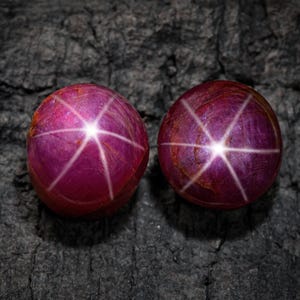 Op de afbeelding: Twee ronde, gepolijste edelstenen met een diepe magenta tint. Elke edelsteen toont een zesstralig sterpatroon dat uitgaat van een centraal lichtpunt. De edelstenen liggen op een donkere, getextureerde ondergrond.