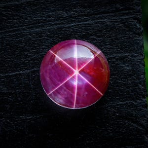 Puede incluir: Una gema de rubí pulida y redonda con un efecto de estrella de seis rayos. La gema es de color rojo púrpura intenso con una superficie brillante y reflectante. El patrón de estrella es de un tono rosa más claro. La gema está sobre un fondo negro.