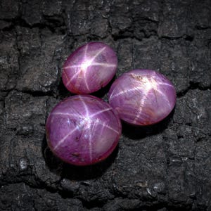 May include: Three oval star ruby gemstones, each displaying a six-ray star pattern. The gems are a deep pinkish-purple hue, set against a dark, textured background.
