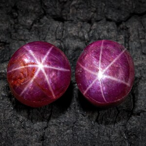 Puede incluir: Dos rubíes estrella redondos y pulidos con un patrón de estrella de seis rayos. Las gemas son de un color rojo púrpura intenso y se asientan sobre una superficie oscura y texturizada. El efecto de estrella se debe a las inclusiones.